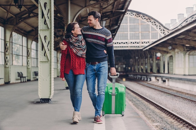 Jovem casal de família se abraça e olha um para o outro com sorriso vestindo jeans e suéteres quentes carregam pose de mala na estação ferroviária se encontram após longa partida namorado e namorada abraçados do lado de fora