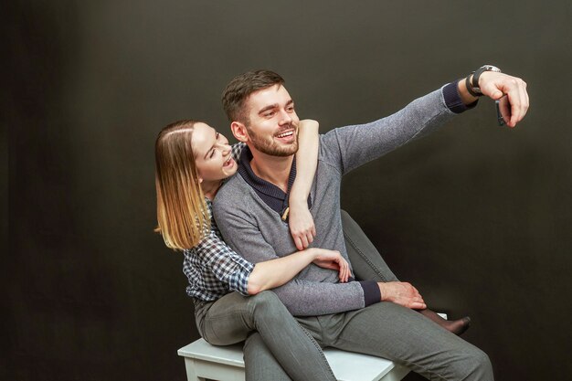 Jovem casal de homem barbudo bonito e mulher loira, sentado contra um fundo preto e fazendo um selfie engraçado. Foto de estúdio