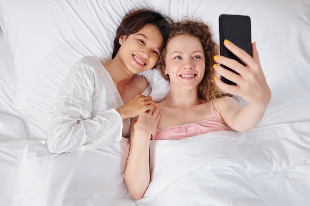 Jovem casal de lésbicas multiétnicas feliz, deitado na cama e tirando uma selfie