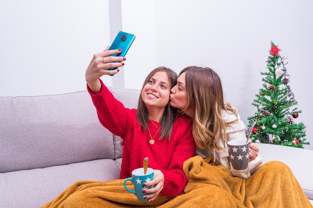 Jovem casal de lésbicas tomando café, tirando fotos e se divertindo perto da árvore de natal. conceito, relaxamento e vida familiar do casal lgbt.