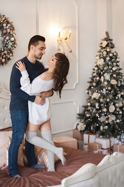 Jovem casal de namorados se abraçando na cama em um quarto ao lado de uma árvore de natal decorada