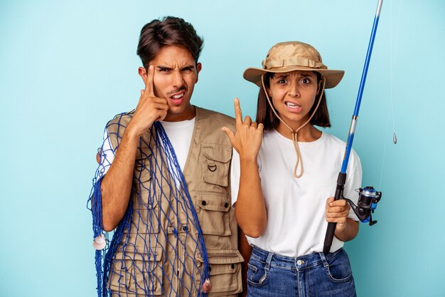 Jovem casal de pescadores de raça mista isolado em um fundo azul, mostrando um gesto de decepção com o dedo indicador.