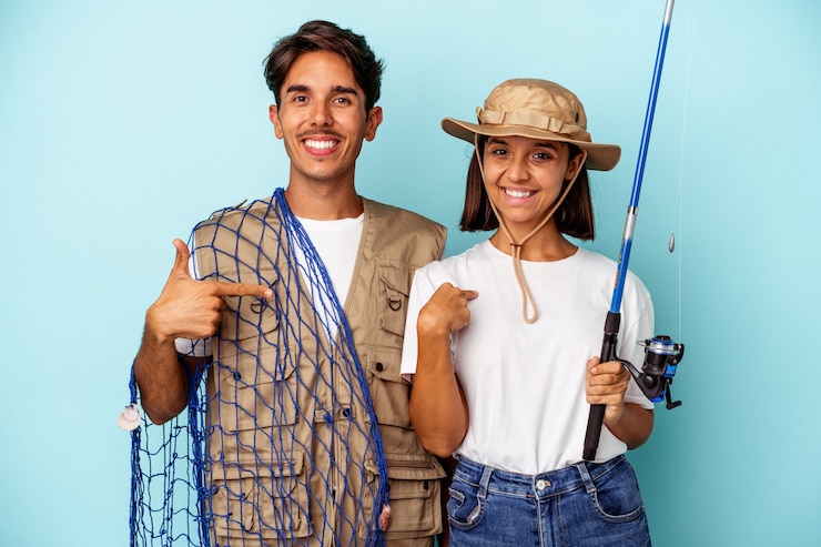 Foto jovem casal de pescadores de raça mista isolado em um fundo azul pessoa apontando com a mão para um espaço de cópia de camisa, orgulhoso e confiante