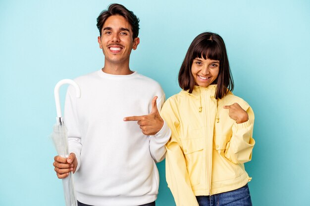 Jovem casal de raça mista segurando guarda-chuva isolado em um fundo azul pessoa apontando com a mão para um espaço de cópia de camisa, orgulhoso e confiante