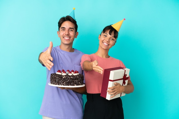 Jovem casal de raça mista segurando um bolo de aniversário e um presente isolado em um fundo azul, apertando as mãos para fechar um bom negócio