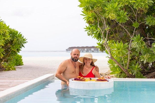 jovem casal de recém-casados passam tempo juntos em lua-de-mel admirando na piscina com servido bandeja flutuante bebidas e lanches em resort de ilha tropical nas Maldivas pequeno-almoço para data romântica luxo