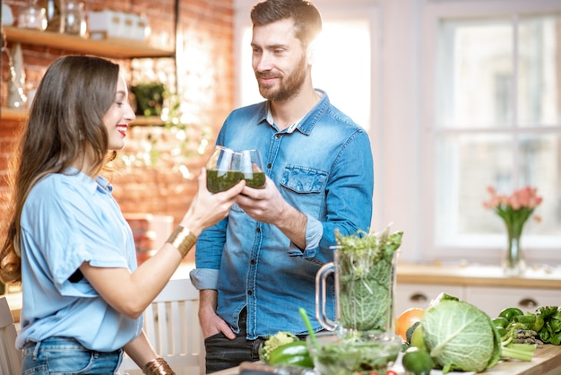 Jovem casal de vegetarianos bebendo smoothie fresco na cozinha com comida verde saudável em casa