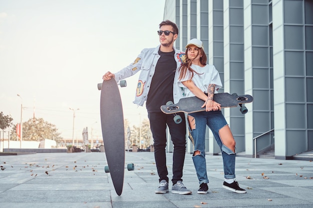 Jovem casal de vestidos com roupas da moda posando com skates perto de arranha-céus.