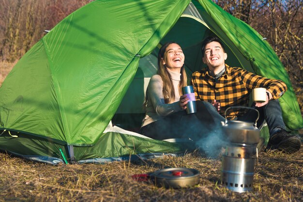 Jovem casal desfrutando ao ar livre, acampar na natureza com tenda bebe chá por uma fogueira