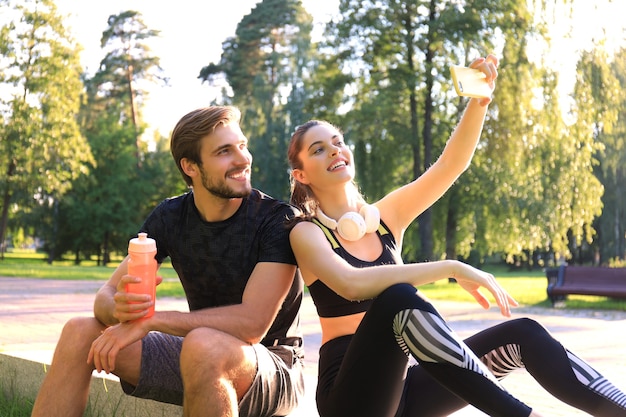 Jovem casal desportivo em roupas esportivas, sentado no parapeito no parque urbano e tomando selfie após o treino em um dia ensolarado de verão.