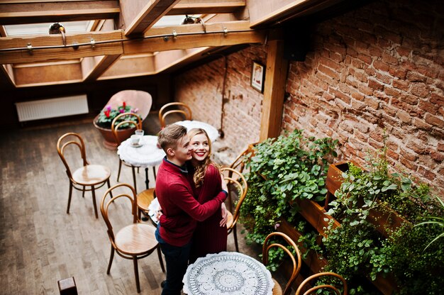 Jovem casal elegante lindo em um vestido vermelho em história de amor no café vintage com grandes janelas no telhado