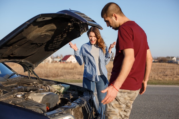 Jovem casal em pé perto de carro quebrado, jura na frente do gancho aberto, macho tenta reparar o veículo e sua esposa grita alto