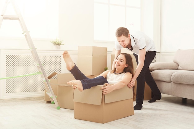 Jovem casal embalando caixas móveis em apartamento novo, se divertindo. homem montando mulher sentada na caixa, copie o espaço