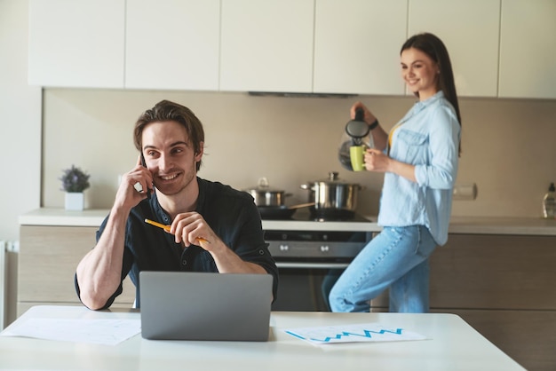 Jovem casal enquanto trabalhava em casa. Marido sentado em uma mesa em sua cozinha usando um laptop enquanto sua esposa está cozinhando em segundo plano.