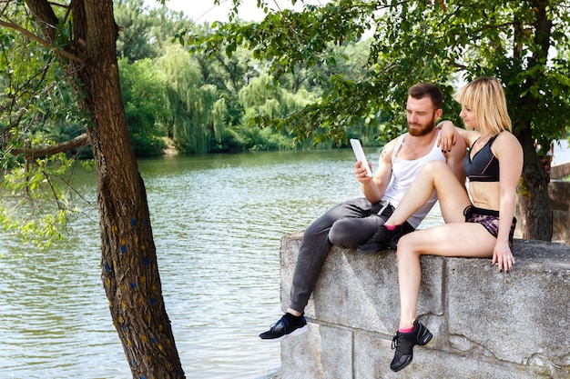 Jovem casal esportiva sentado em um parque perto de um rio, usar internet no tablet PC branco