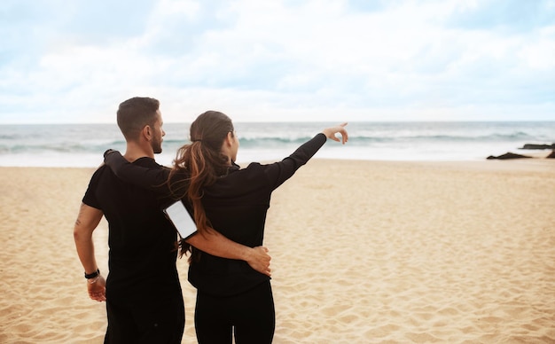 Jovem casal esportivo malhando juntos na praia à beira-mar descansando depois de treinar abraçando a mulher apontando para fora