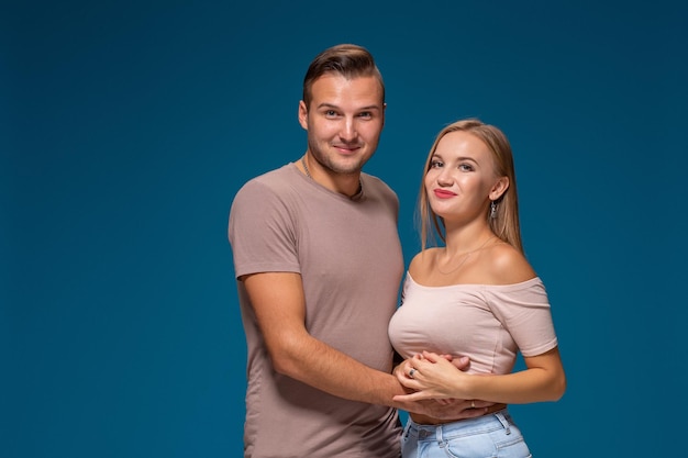 Jovem casal está abraçando em um fundo azul no estúdio. Eles vestem camisetas, jeans e sorriem.