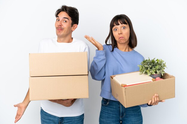 Jovem casal fazendo um movimento enquanto pega uma caixa cheia de coisas isoladas no fundo branco, fazendo gesto de dúvidas enquanto levanta os ombros
