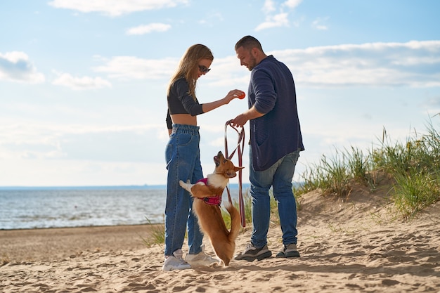 Jovem casal feliz brincando com o cachorro na praia. Homem segurando cachorro Corgi na coleira