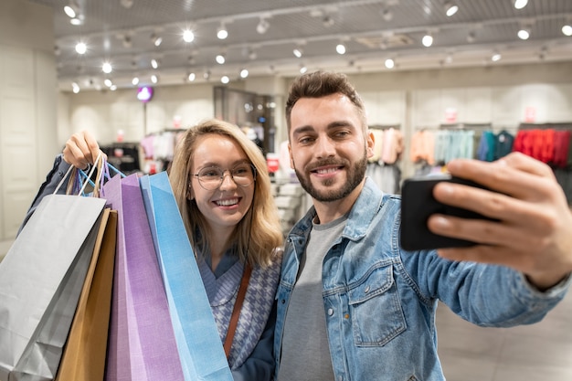 Jovem casal feliz com sacolas de compras fazendo selfie retrato no celular na loja