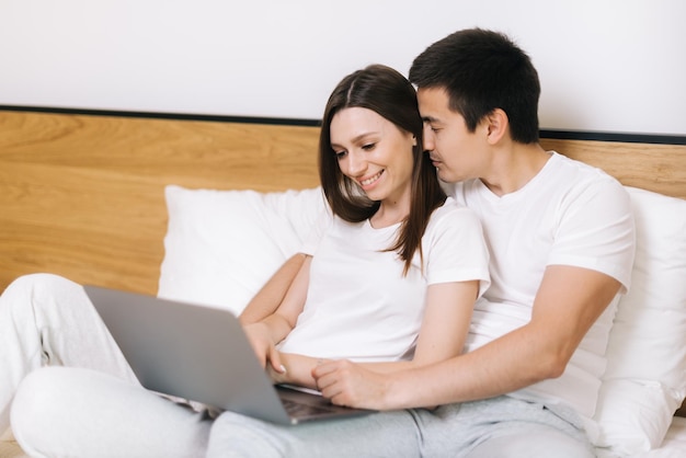 Jovem casal feliz de homem e mulher está deitado na cama e assistindo filme no laptop. Jovem casal deitado na cama em casa e usando o laptop. Casal feliz está descansando na cama antes de ir para a cama