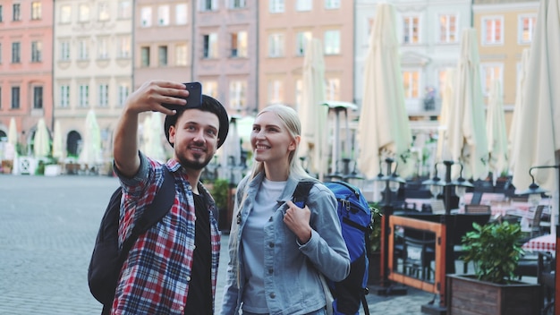 Jovem casal feliz de turistas fazendo selfie em smartphone no centro da cidade. Eles têm bolsas de turistas.