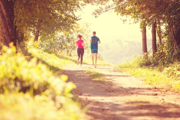 Foto jovem casal feliz desfrutando de um estilo de vida saudável enquanto faz jogging em uma estrada rural através da bela floresta ensolarada, exercício e conceito de fitness