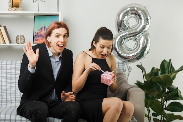 Jovem casal feliz dia da mulher segurando e abrindo um presente sentado no sofá da sala