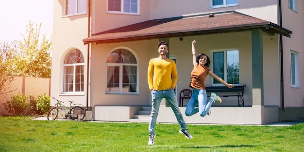 Jovem casal feliz e animado ou esposa e marido apaixonados pulando na frente da nova casa grande enquanto comemoram esta compra