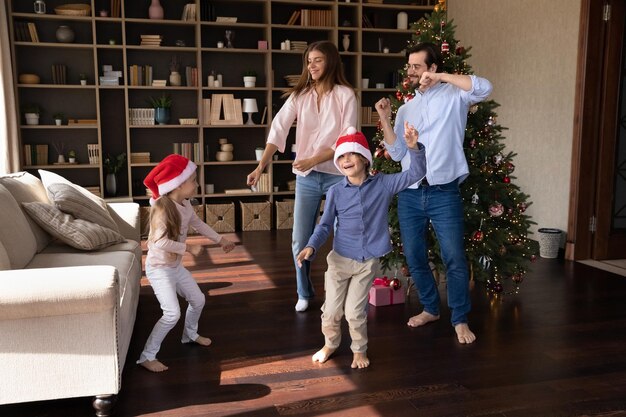 Jovem casal feliz e ativo pais dançando com música festiva de Natal com crianças pequenas bonitas irmãos descalços no chão de madeira quente perto de árvore decorada feriados de Ano Novo conceito de celebração em família