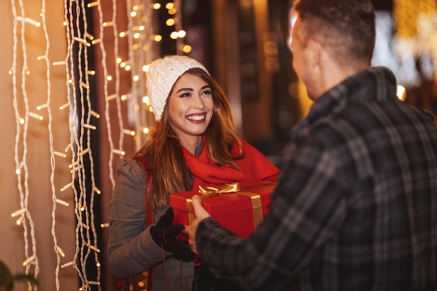Jovem casal feliz está andando pela rua da cidade com férias brilhantes no fundo. Homem sorridente está dando um presente para sua namorada feliz em uma noite de Natal.