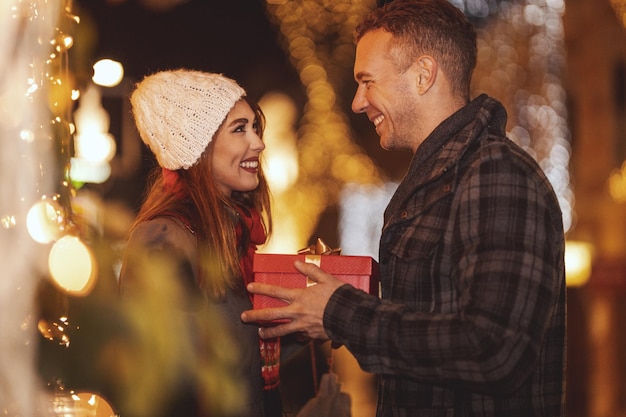 Jovem casal feliz está andando pela rua da cidade com férias brilhantes no fundo. Homem sorridente está dando um presente para sua namorada feliz em uma noite de Natal.