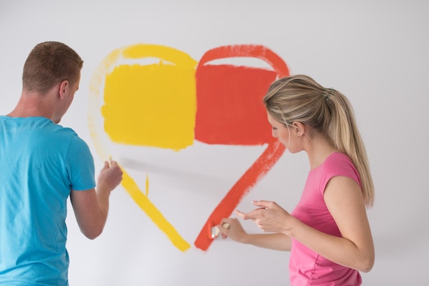 Jovem casal feliz está pintando um coração na parede enquanto faz reparos em casa.