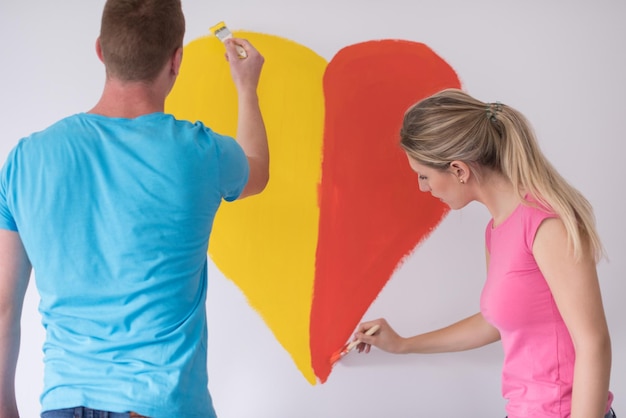 Jovem casal feliz está pintando um coração na parede enquanto faz reparos em casa.