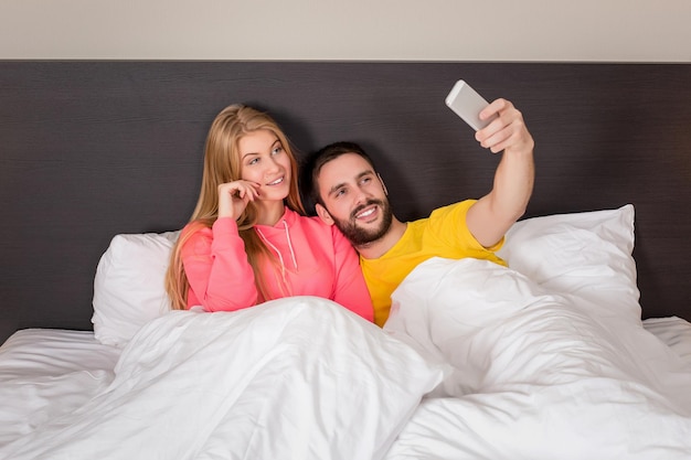 Jovem casal feliz na cama fazendo selfie com câmera de telefone. conceito sobre tecnologia e pessoas