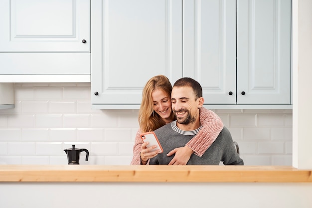 Jovem casal feliz na cozinha de casa, usando telefone celular