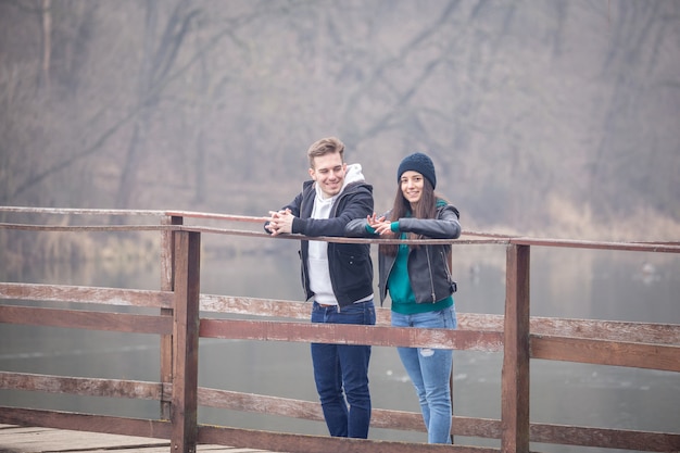 Jovem casal feliz parado em uma velha ponte de madeira em um dia nublado de inverno no Lago Gebart