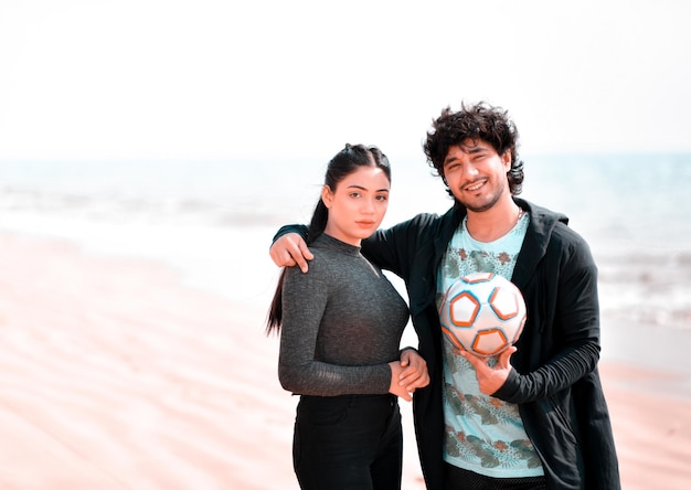jovem casal feliz pose frontal segurando futebol na praia modelo paquistanês indiano