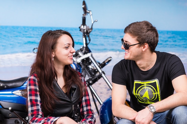 Jovem casal feliz relaxando à beira-mar, sentado em frente a sua motocicleta azul, enquanto riem e conversam sob o sol de verão