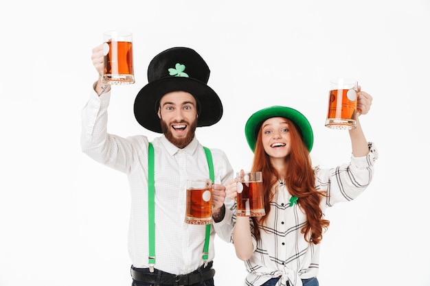 Jovem casal feliz vestindo fantasias, comemorando o dia de St Patrick isolado em uma parede branca, bebendo cerveja