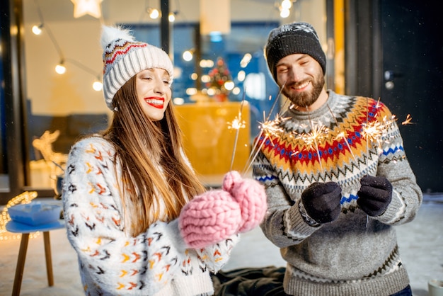 Jovem casal feliz vestindo suéteres comemorando as férias de inverno com uma fogueira de bengala em frente a uma linda casa decorada