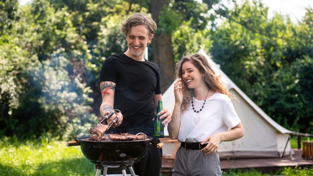 Jovem casal fritando carne na grelha e bebendo cerveja, rindo. Vegetação ao redor. Glamping