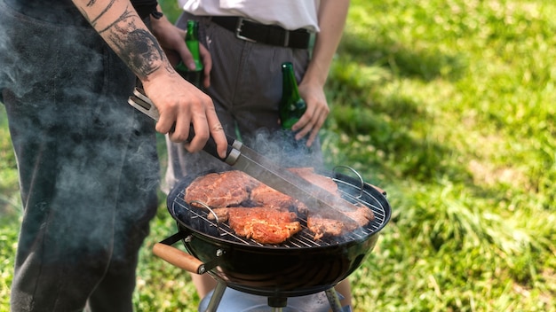 Jovem casal fritando carne na grelha e bebendo cerveja. Vegetação ao redor. Glamping