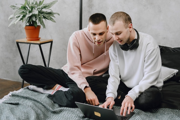 Jovem casal gay sentado na cama usando um laptop, usando fones de ouvido, ouvindo música juntos, se abraçando