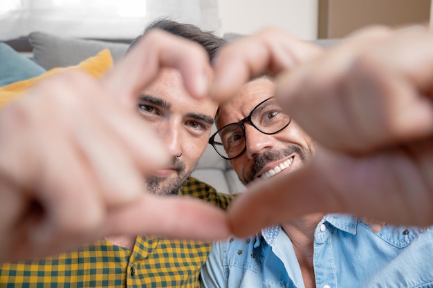 Jovem casal gay sorrindo confiante fazendo símbolo de coração com as mãos em casa