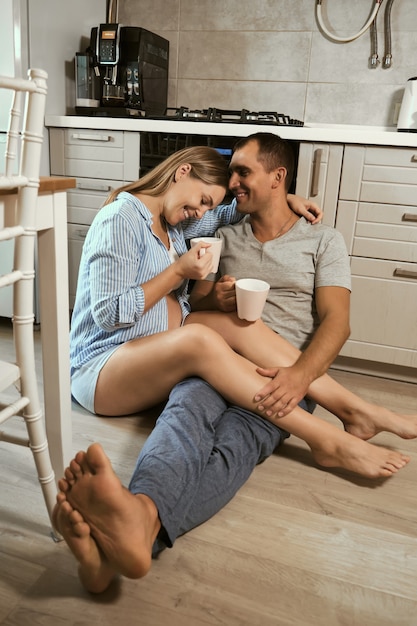 Jovem casal grávida na cozinha no chão, abraço, ria com xícaras de chá e regozijando-se na expectativa do bebê. Vida nova do conceito, jovem família.