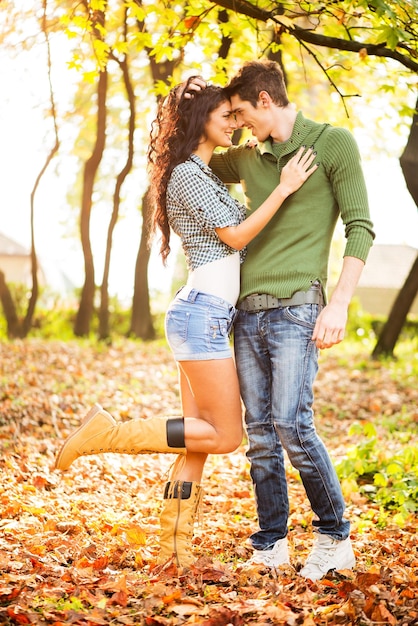 Foto jovem casal heterossexual apaixonado no parque, de pé sobre folhas caídas, inclinando a cabeça um para o outro, olhando um para o outro e sorrindo.