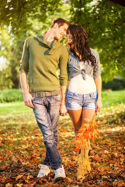 Jovem casal heterossexual no parque, andando sobre folhas caídas, de mãos dadas e olhando um para o outro com um sorriso.