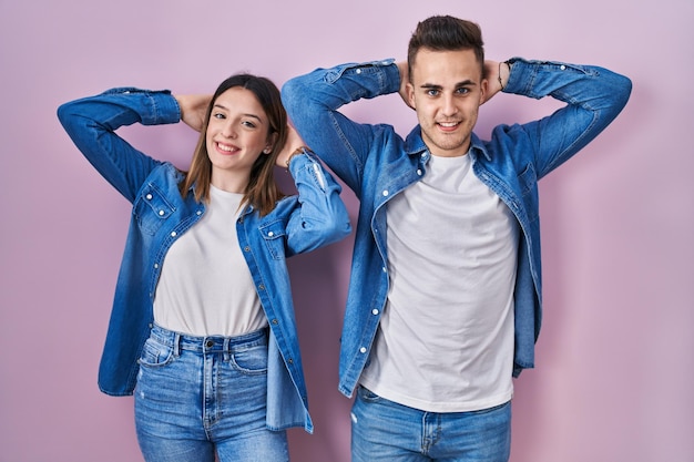 Jovem casal hispânico de pé sobre fundo rosa relaxando e esticando braços e mãos atrás da cabeça e pescoço sorrindo feliz