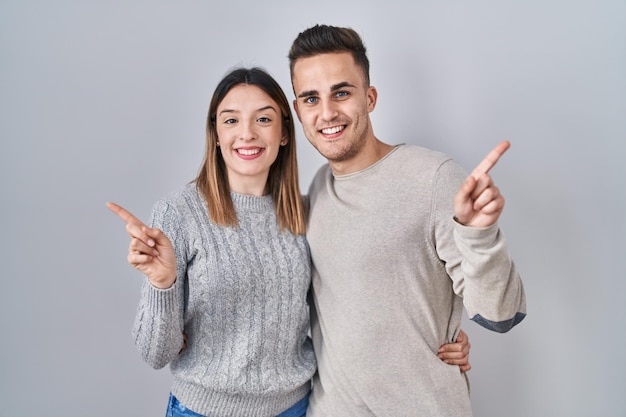 Jovem casal hispânico em pé sobre fundo branco com um grande sorriso no rosto apontando com o dedo da mão para o lado olhando para a câmera